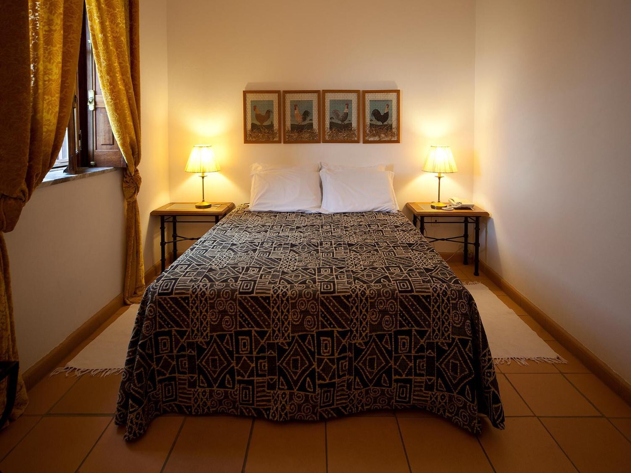 Een slaapkamer met een bedovertrek met patroon, twee bedlampen, een raam met gordijnen en drie ingelijste foto's aan de muur.