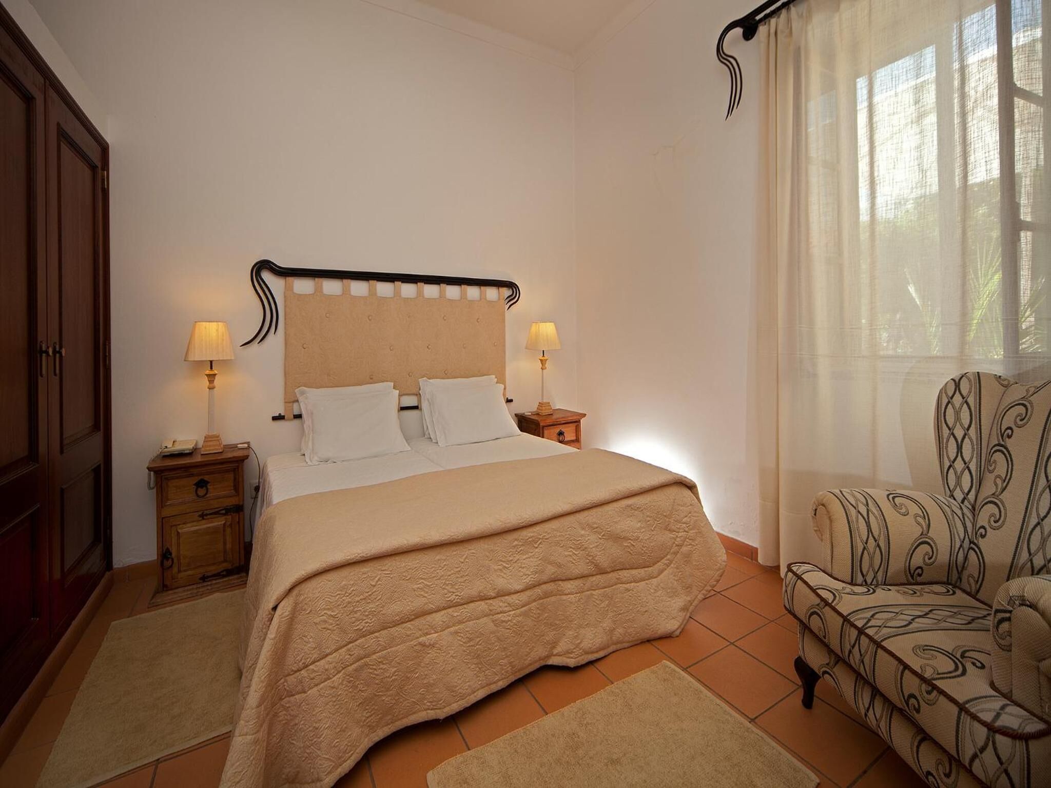 Un dormitorio con cama, dos mesitas de noche con lámparas, una silla y una ventana con cortinas.