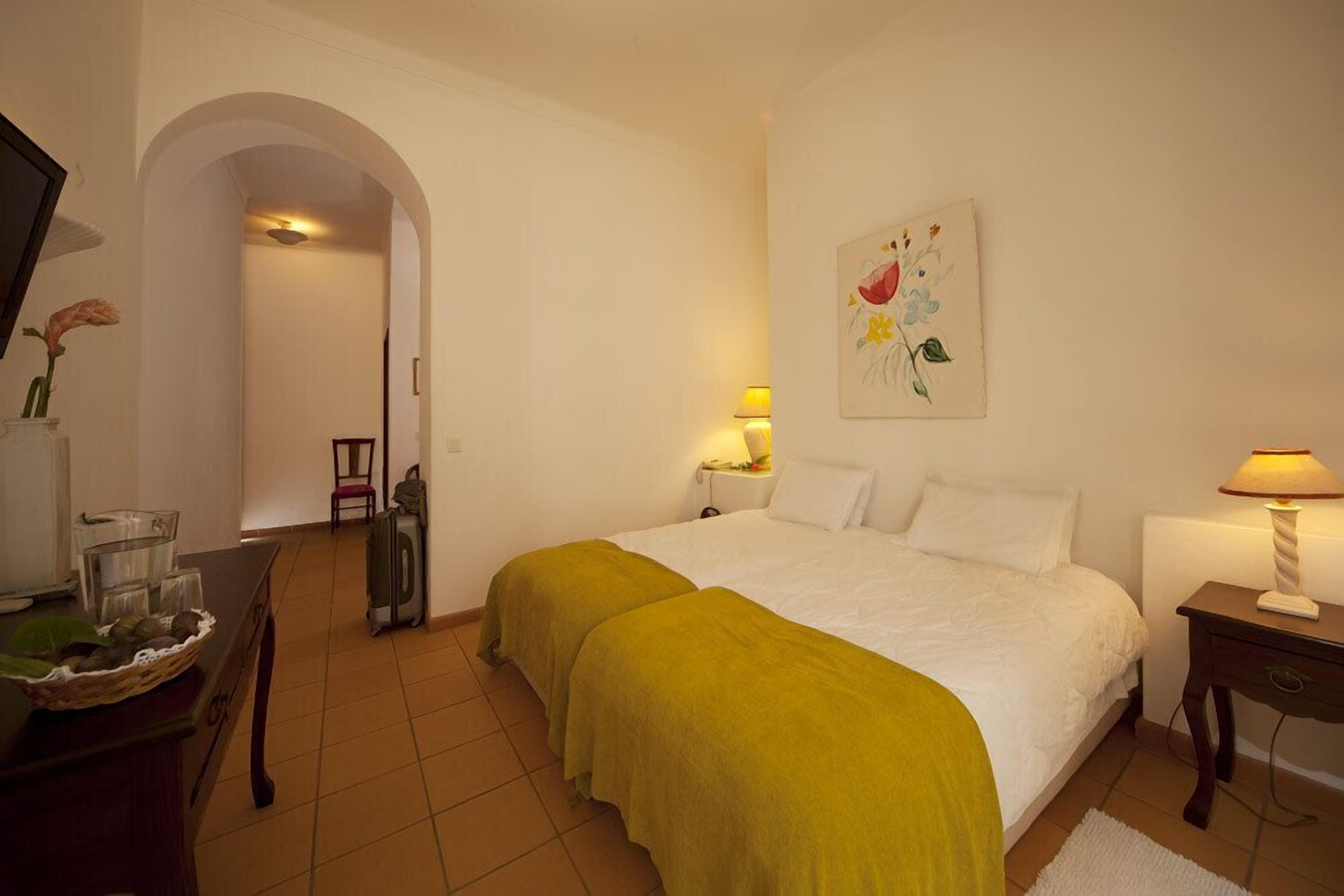 A hotel room with a bed, two bedside lamps, a desk with a lamp, a chair, a TV, and a painting on the wall.