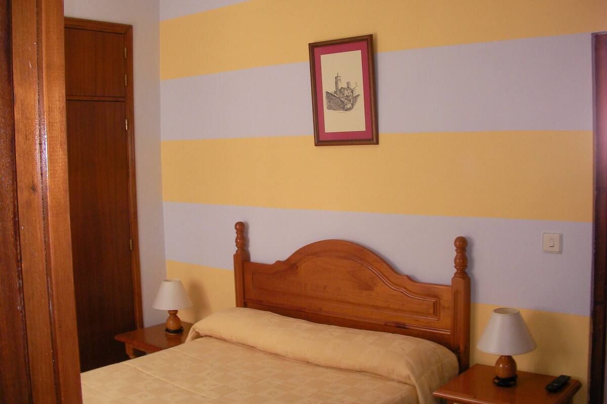 Un dormitorio con cama de madera, dos mesitas de noche con lámparas, un cuadro enmarcado en la pared y una puerta a la izquierda.