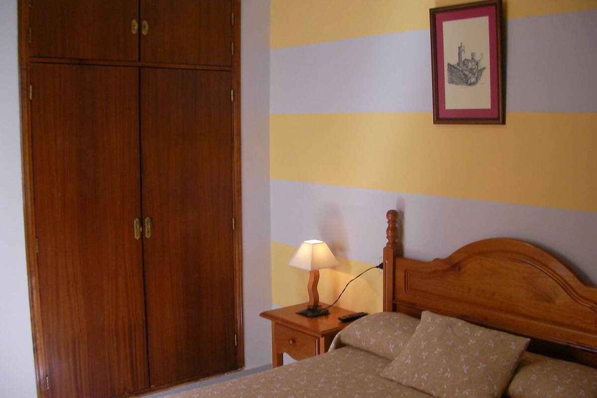 Un dormitorio con un armario de madera, una mesita de noche con lámpara y una cama con ropa de cama estampada.