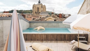 Una piscina en la azotea con una gran decoración inflable de langosta, sillones de descanso y una vista de una iglesia histórica al fondo.