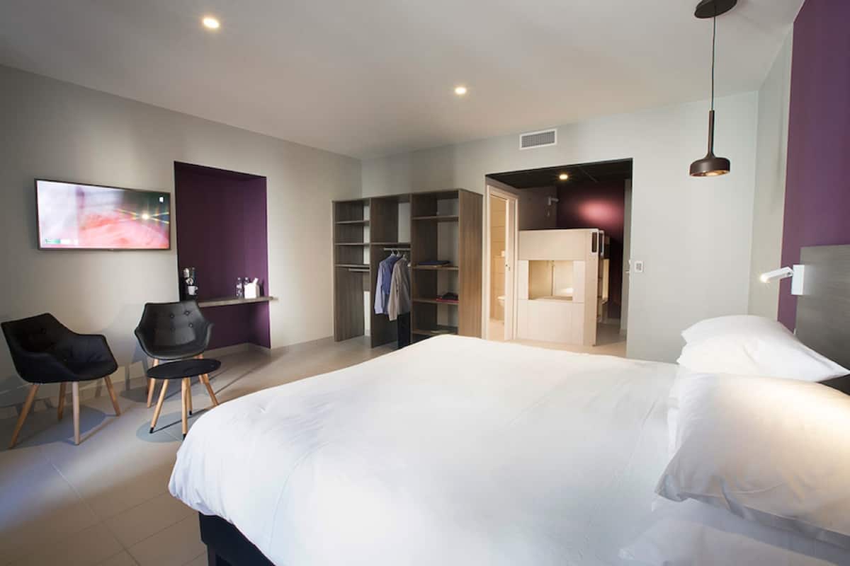 A modern hotel room with a large bed, a desk, a chair, a wardrobe, and a television.