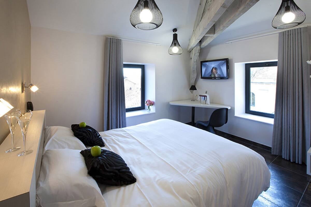 A hotel room with a large bed, a desk, two windows, and a television.