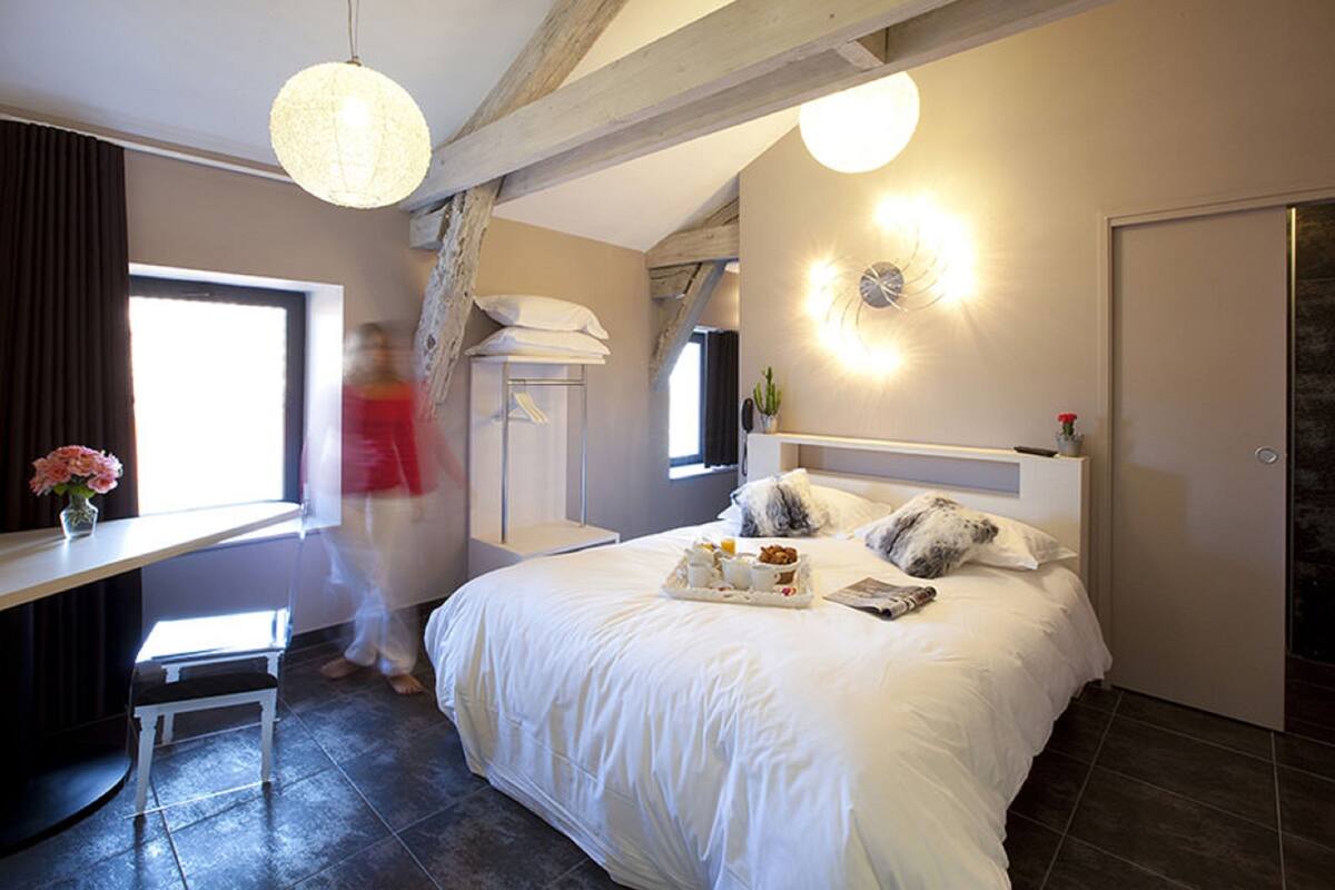 A bedroom with a large bed, a desk, and a chair. There is a window with curtains, a wall clock, and a door.