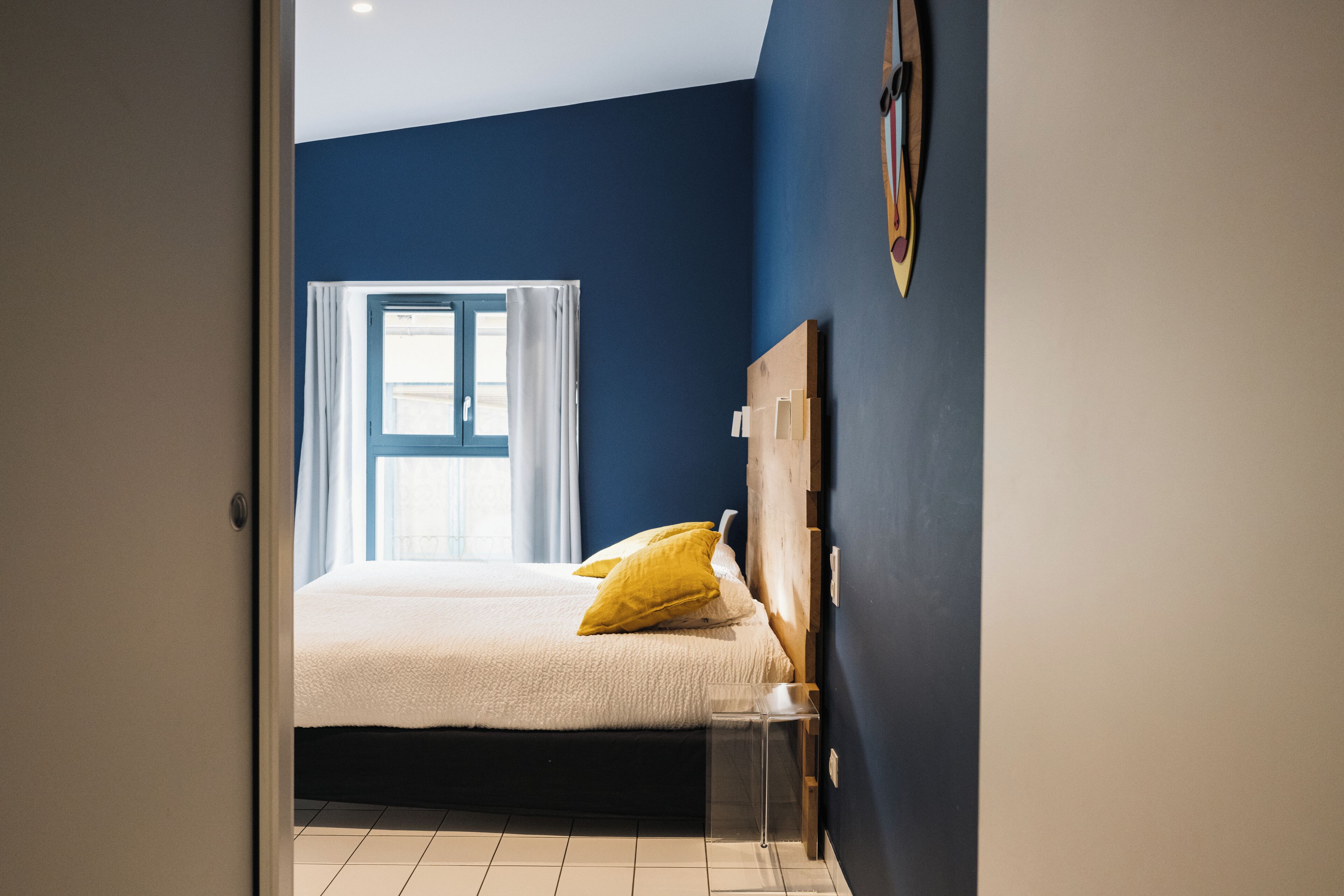A bedroom with a bed, a window with curtains, a wooden headboard, and a small bedside table.
