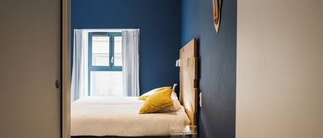 A bedroom with a bed, a window with curtains, a wooden headboard, and a small bedside table.