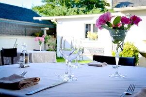 A table set for dining outdoors with a clear glass vase containing pink flowers, a water pitcher, and wine glasses.
