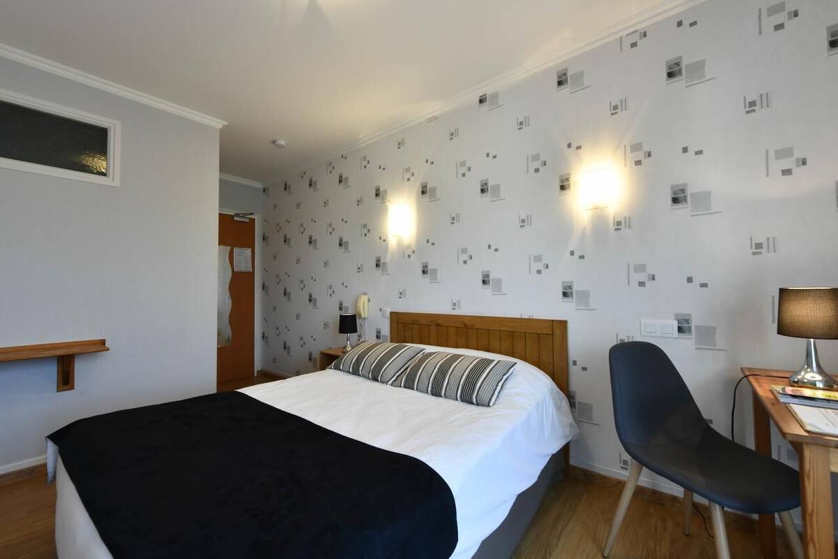 A hotel room with a bed, a chair, a desk, and a wall with patterned wallpaper.
