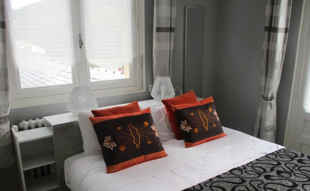 Habitación de hotel con cama, dos almohadas decoradas con motivos florales, mesita de noche, lámpara y ventana con cortinas.