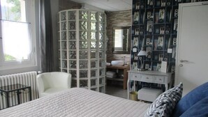 A bedroom with a bed, a white chair, a vanity with a mirror, and a wall with framed pictures. - L'Ayguelade (Bielle)
