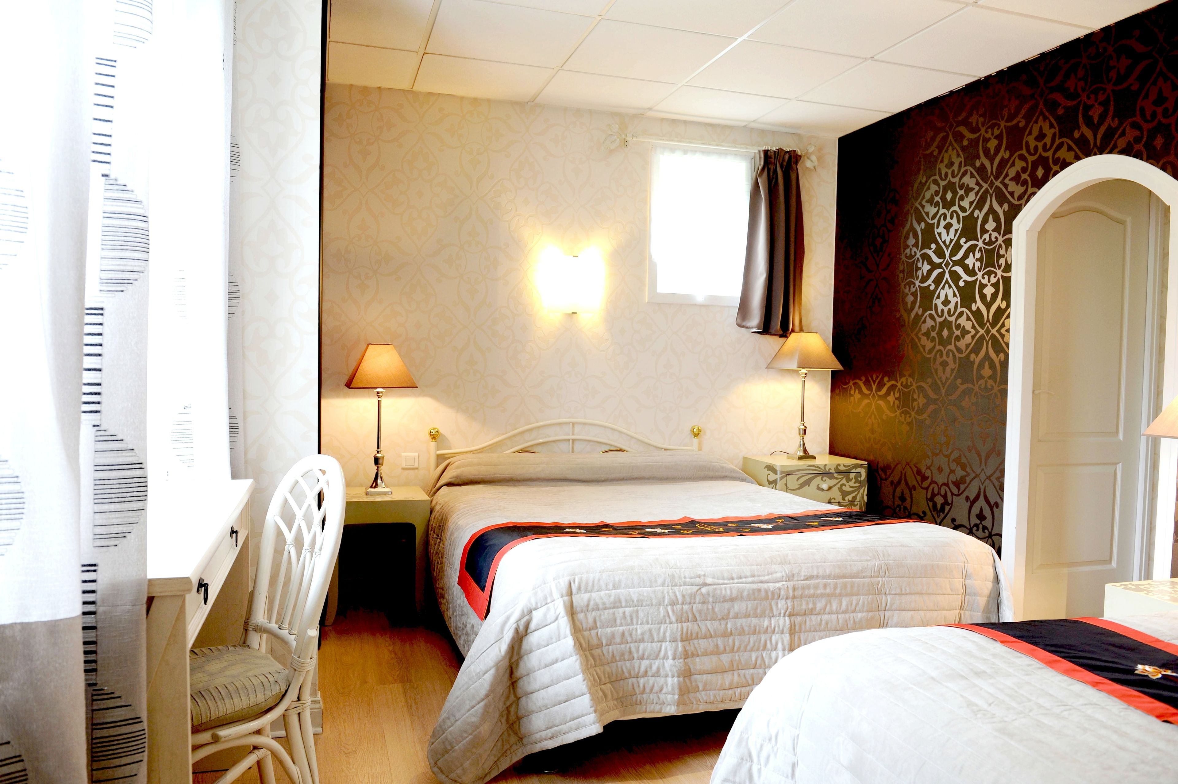 Ein Hotelzimmer mit zwei Betten, einem Schreibtisch und einem Sessel. Zwei Nachttische mit Lampen sind vorhanden.