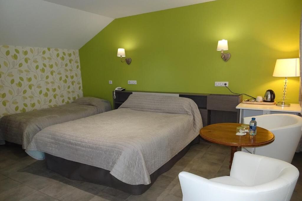 A hotel room with two beds, a small round table with a chair, a desk with a lamp, and two wall-mounted lamps.