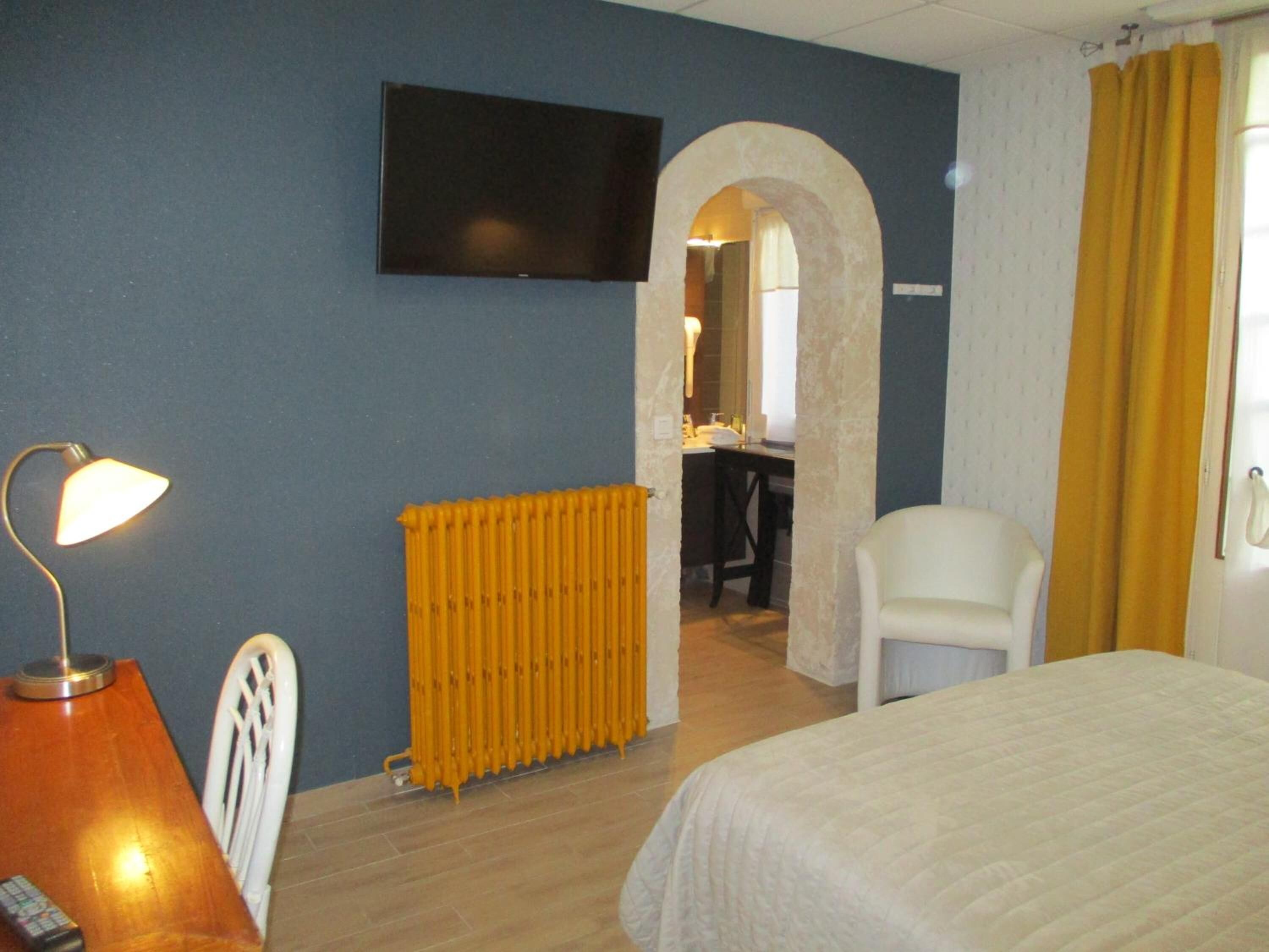 Habitación de hotel con cama, escritorio con lámpara, silla, radiador y televisor en la pared.