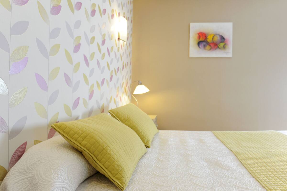 A neatly made bed with two pillows, a wall with a leaf pattern, a wall clock, and a framed artwork.