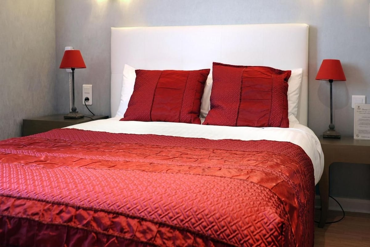 A neatly made bed with a red quilt and pillows, two bedside lamps, and a nightstand with a lamp.
