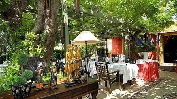 An outdoor dining area with tables and chairs, surrounded by greenery and decorative statues.