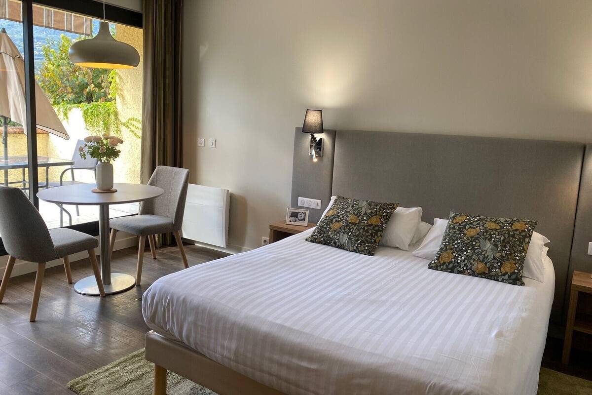 A hotel room with a large bed, a round dining table, two chairs, a small table with a vase of flowers, a wall-mounted lamp, and a view of greenery outside.