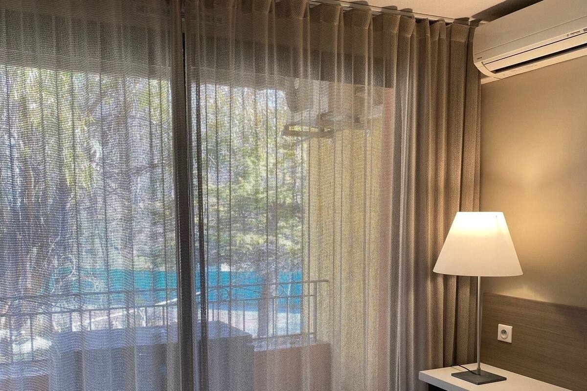 A hotel room with a bed, a desk with a lamp, and sheer curtains allowing natural light.