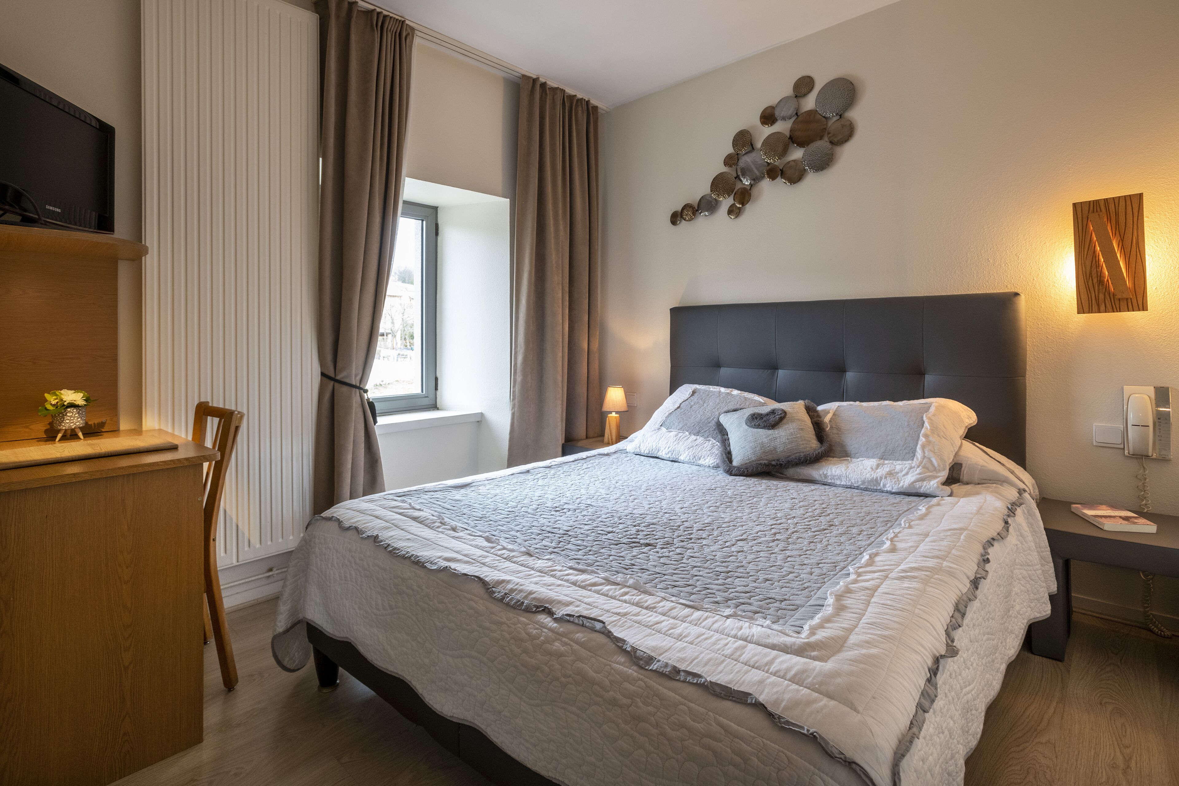 A bedroom with a bed, a desk, a chair, a TV, and a window with curtains.