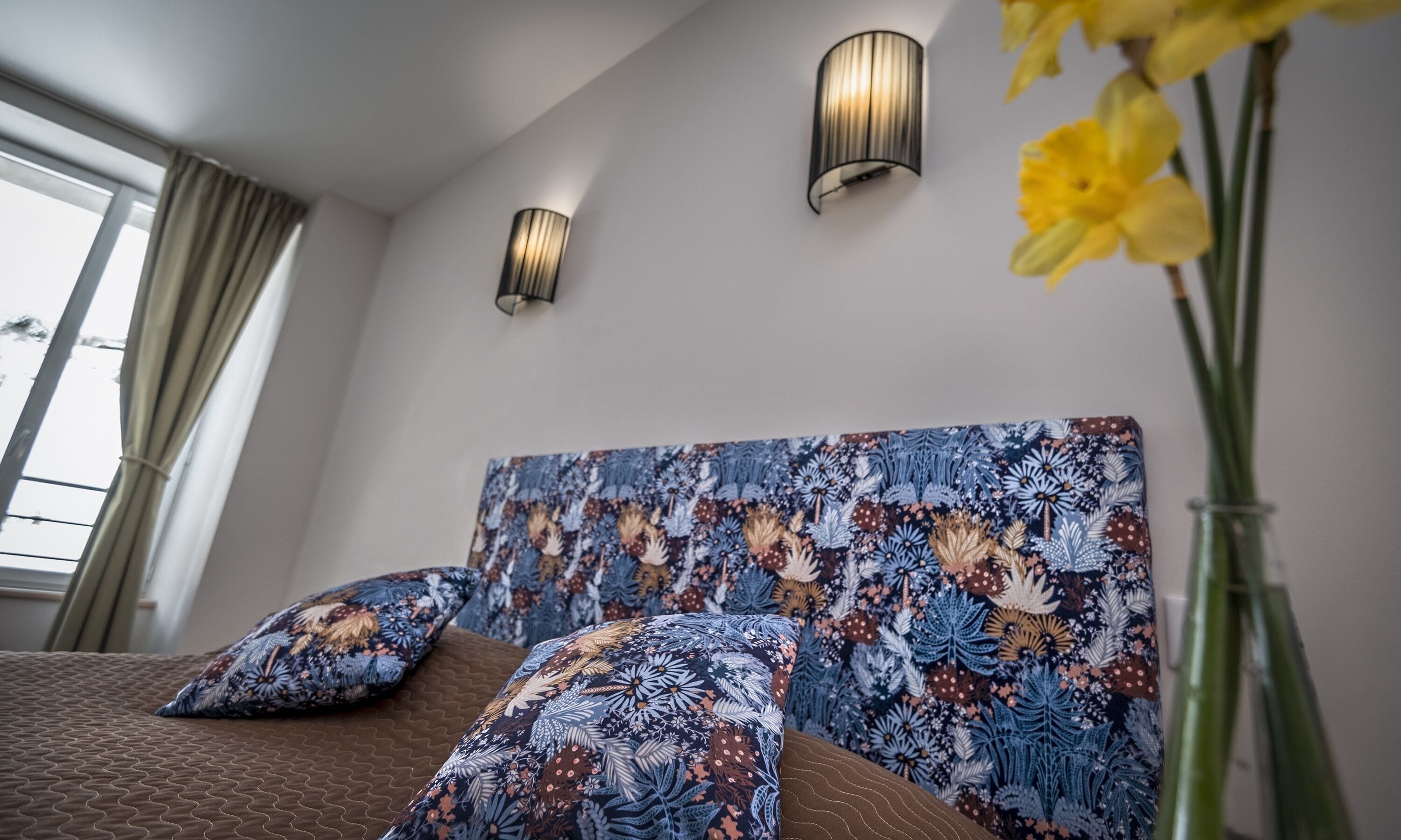 Une chambre à coucher avec un lit, deux lampes fixées au mur et une tête de lit à motifs floraux.