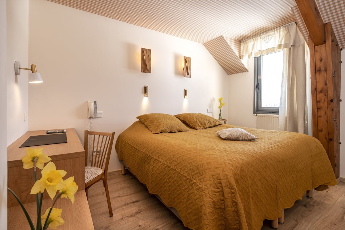 Un dormitorio con cama, escritorio y silla. Hay luces fijadas a la pared y una ventana con cortinas.