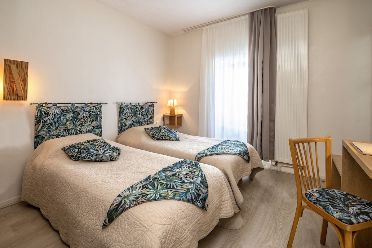 Habitación de hotel con dos camas, una silla de madera, un escritorio y una ventana con cortinas.