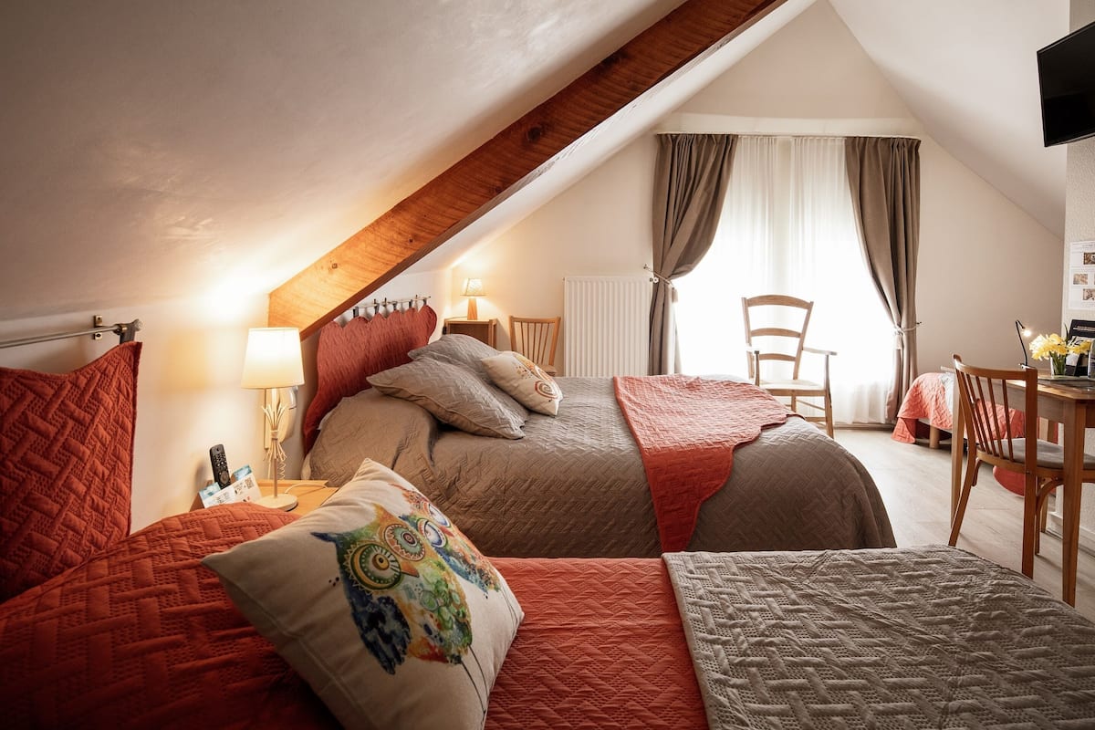 Un acogedor dormitorio en el ático con techo inclinado, cabecera de madera, cama con muchas almohadas, mesita de noche con lámpara, una silla y una mesa con un jarrón de flores.