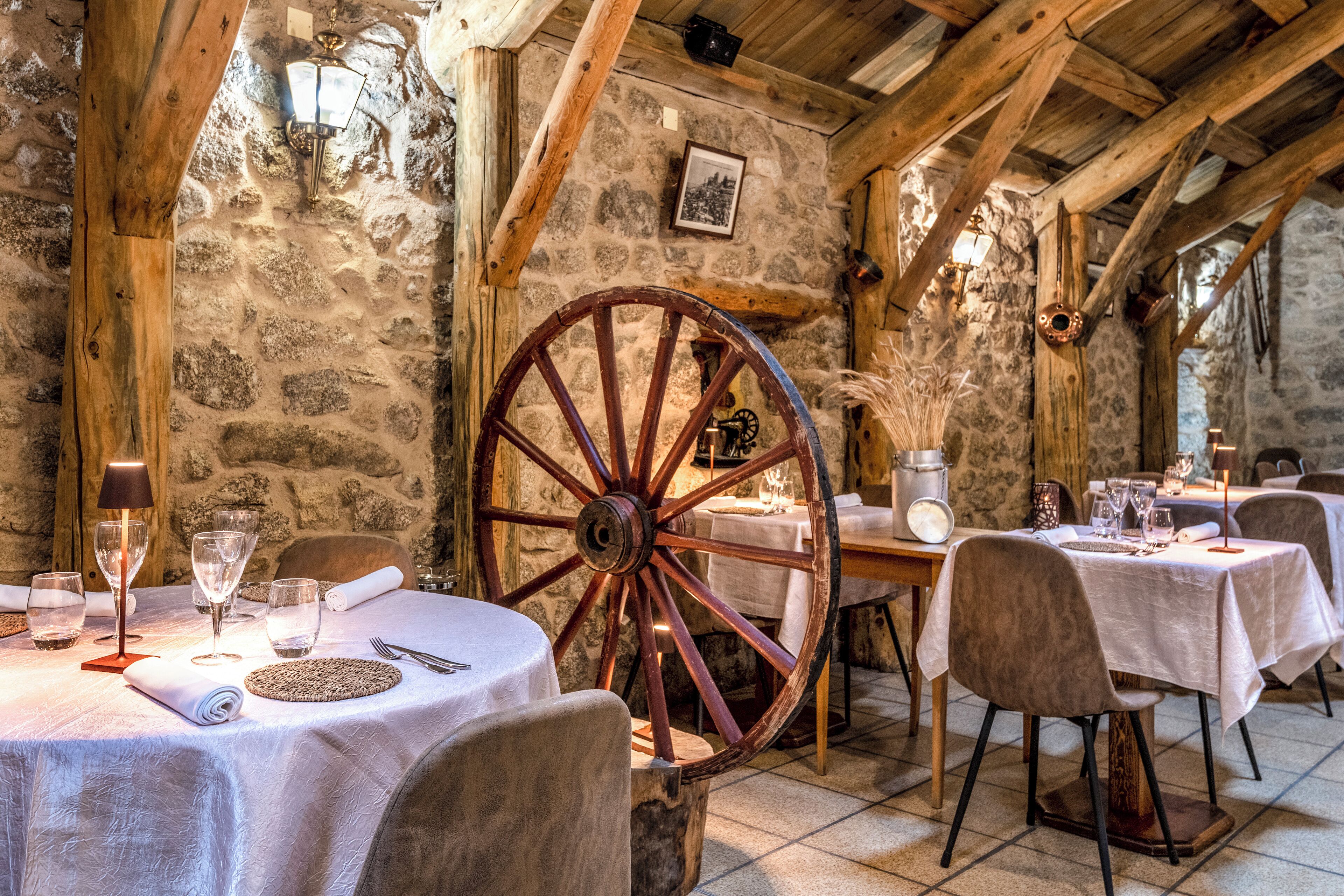 Un comedor rústico con paredes de piedra, vigas de madera y una gran rueda de madera como elemento central. Las mesas están dispuestas con manteles blancos, copas de vino y cubiertos.