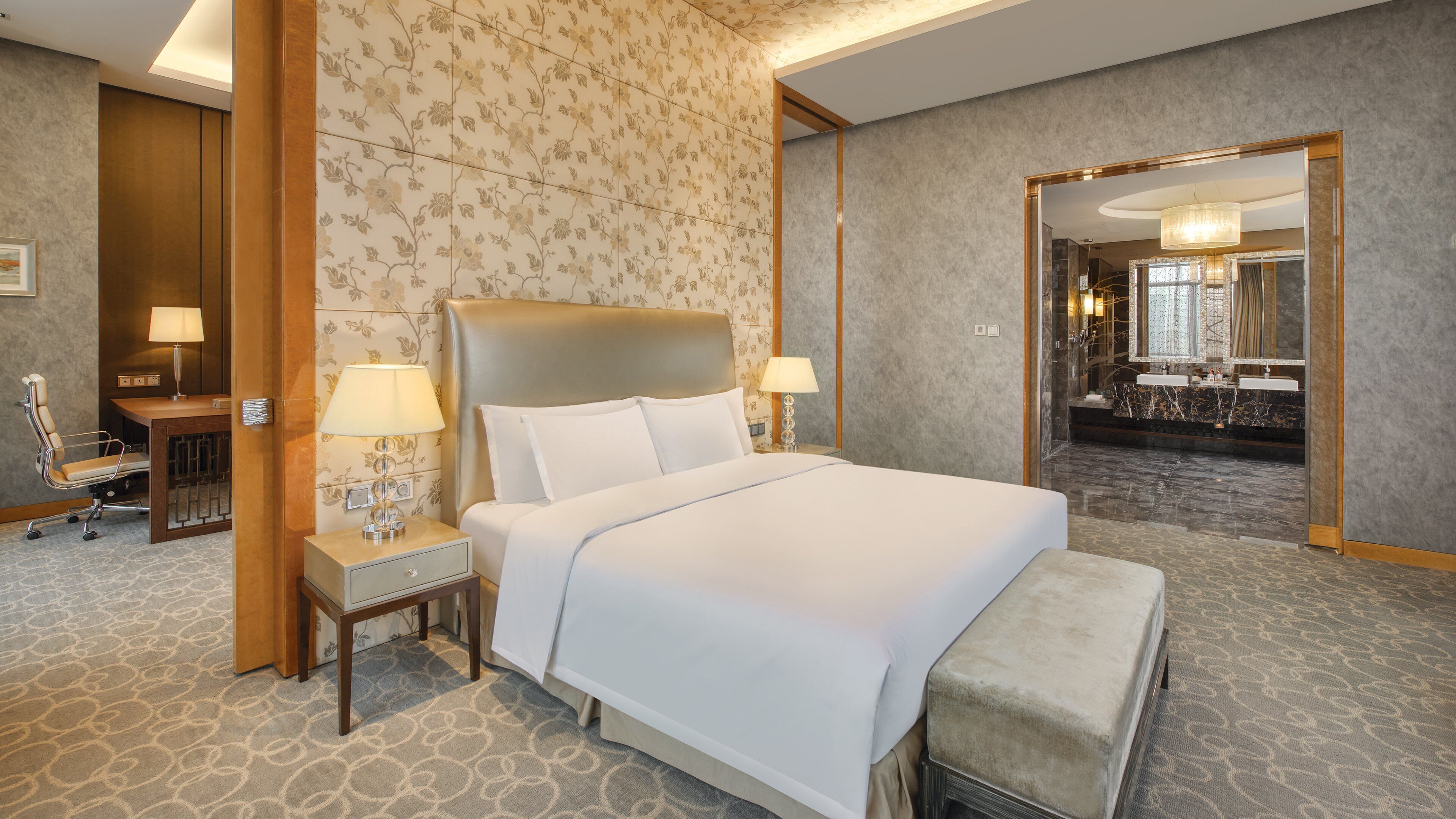 Ein Hotelzimmer mit einem großen Bett, Nachttischlampen, einem Schreibtisch mit Stuhl und einem durchgehenden Türspalte sichtbaren Badezimmer.