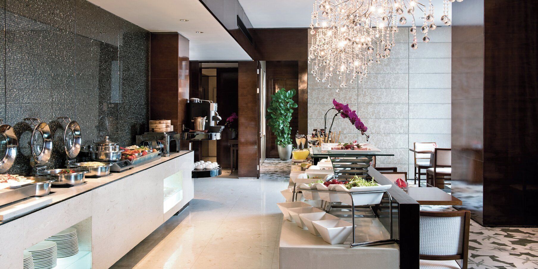 A modern hotel lobby with a buffet area, a dining table with chairs, and a decorative chandelier.