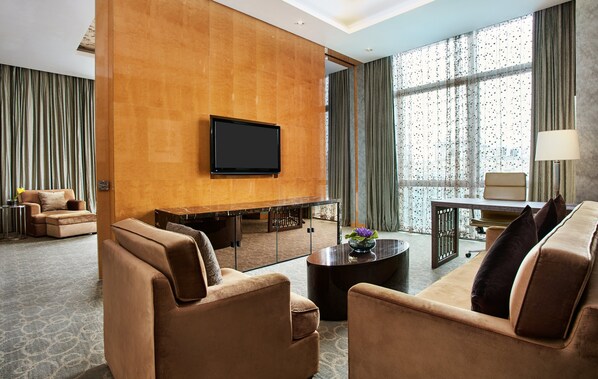 Habitación de hotel moderna con televisor de pantalla plana en pared de madera, un conjunto de sofás, una mesa de centro con un jarrón y vistas amplias a través de ventanas con cortinas.
