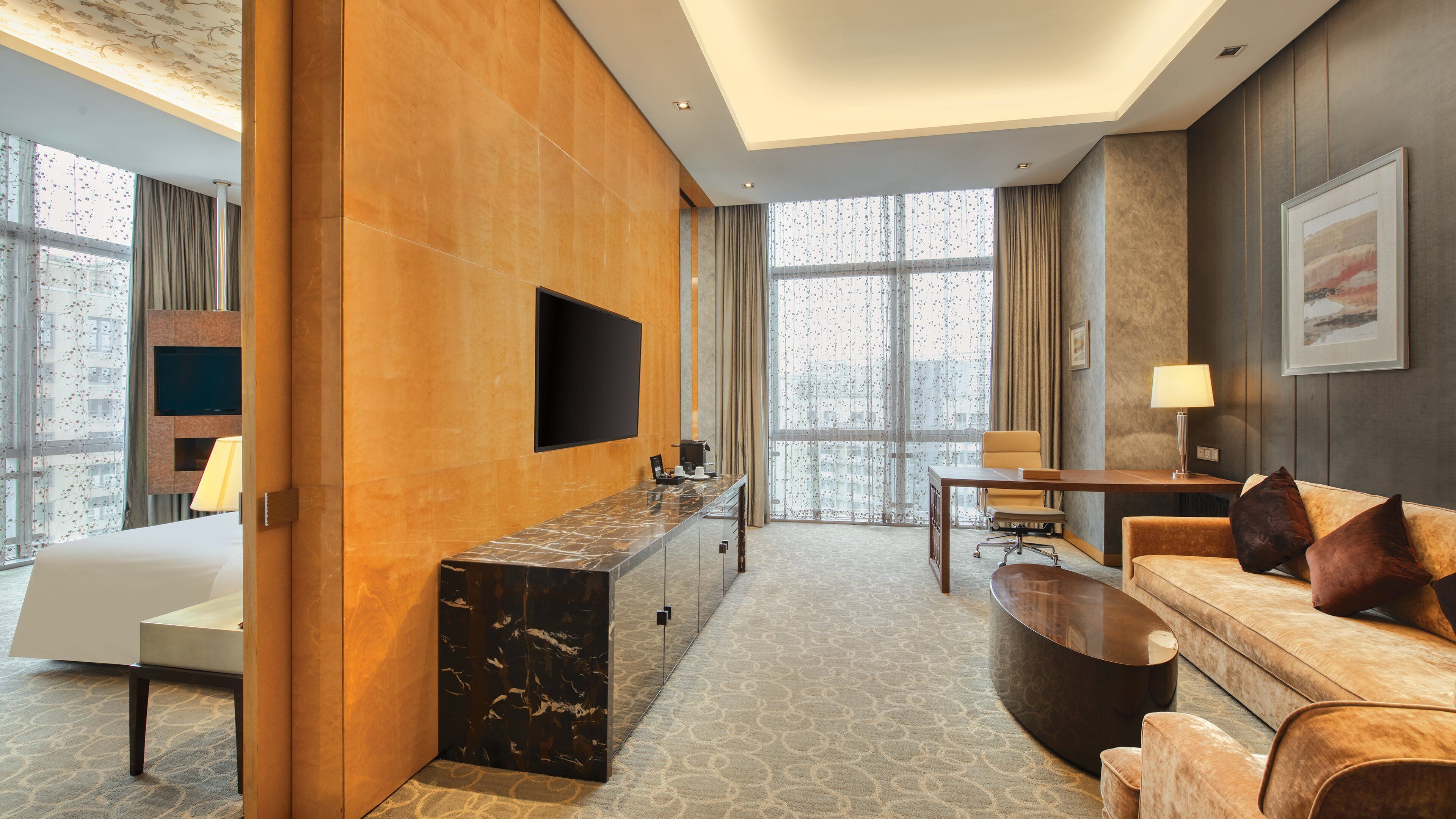Habitación de hotel con un mostrador de recepción de mármol, un televisor de pantalla plana en la pared, un sofá con cojines, una mesita de noche con lámpara y un ventanal con cortinas.