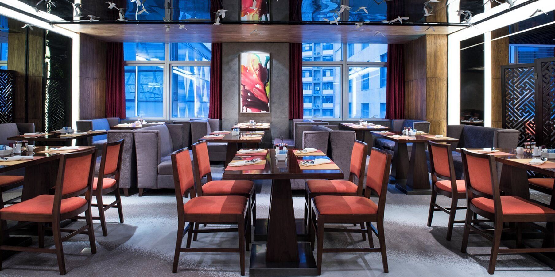 A modern restaurant with multiple dining tables and chairs, large windows with a view of buildings, and decorative wall art.