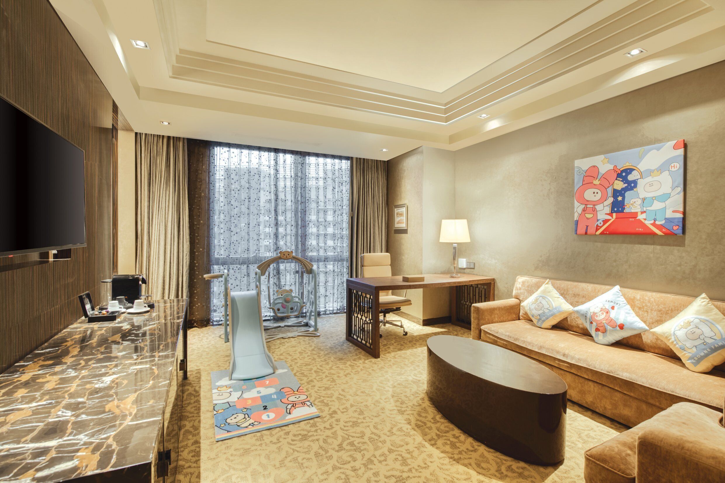 Quarto de hotel com televisão, sofá, mesa de centro, escrivaninha e uma janela grande com cortinas.