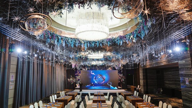 Une salle de conférence dotée d’une scène, de rangées de tables et d’un lustre.