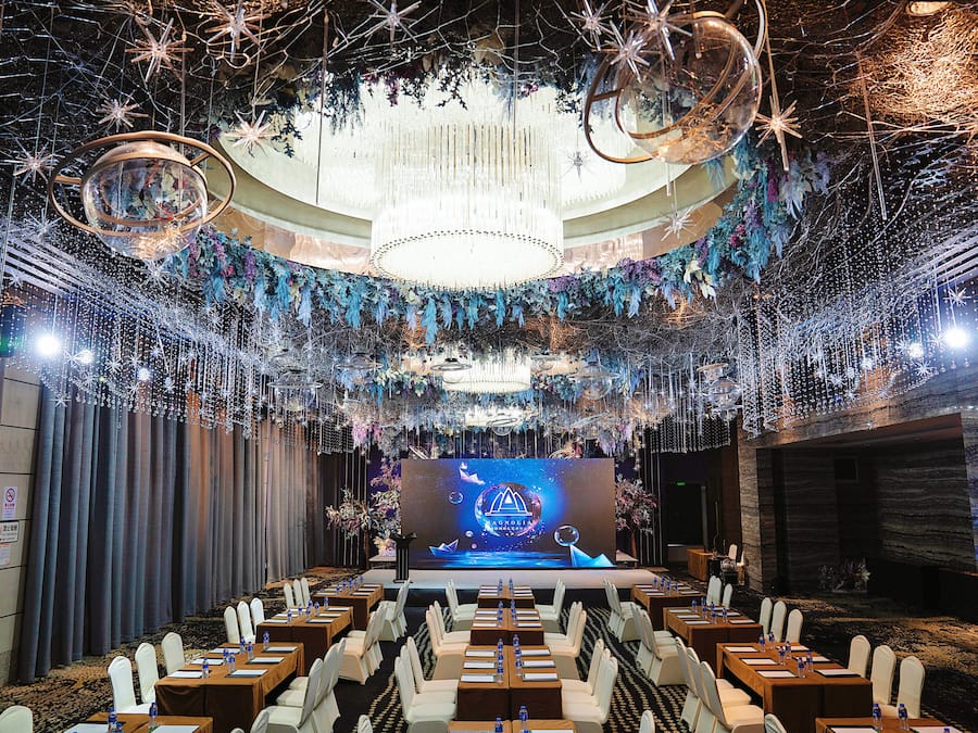 Une salle de conférence dotée d’une scène, de rangées de tables et d’un lustre.