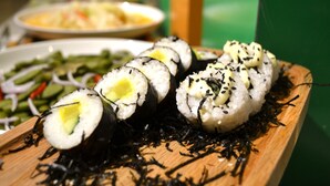 Des rouleaux de sushi présentés sur une planche en bois, garnis d’algues et de graines de sésame, accompagnés d’un bol de légumes verts.