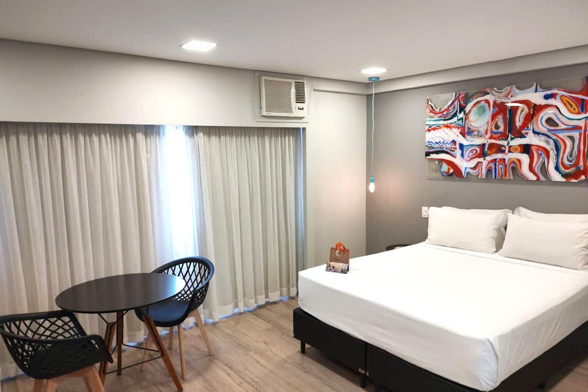 A hotel room with a bed, a table with chairs, a large abstract painting on the wall, and a window with curtains.