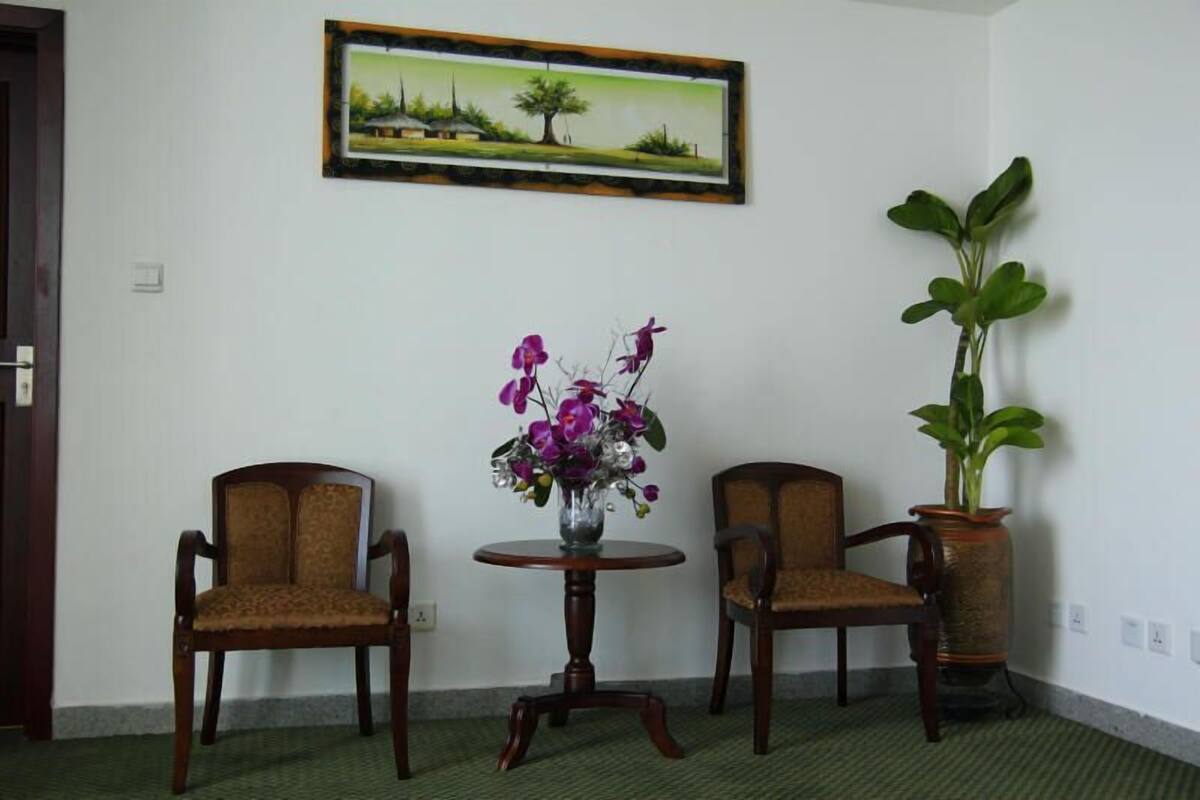 A room with two wooden chairs, a small round table with a vase of flowers, and a large potted plant.