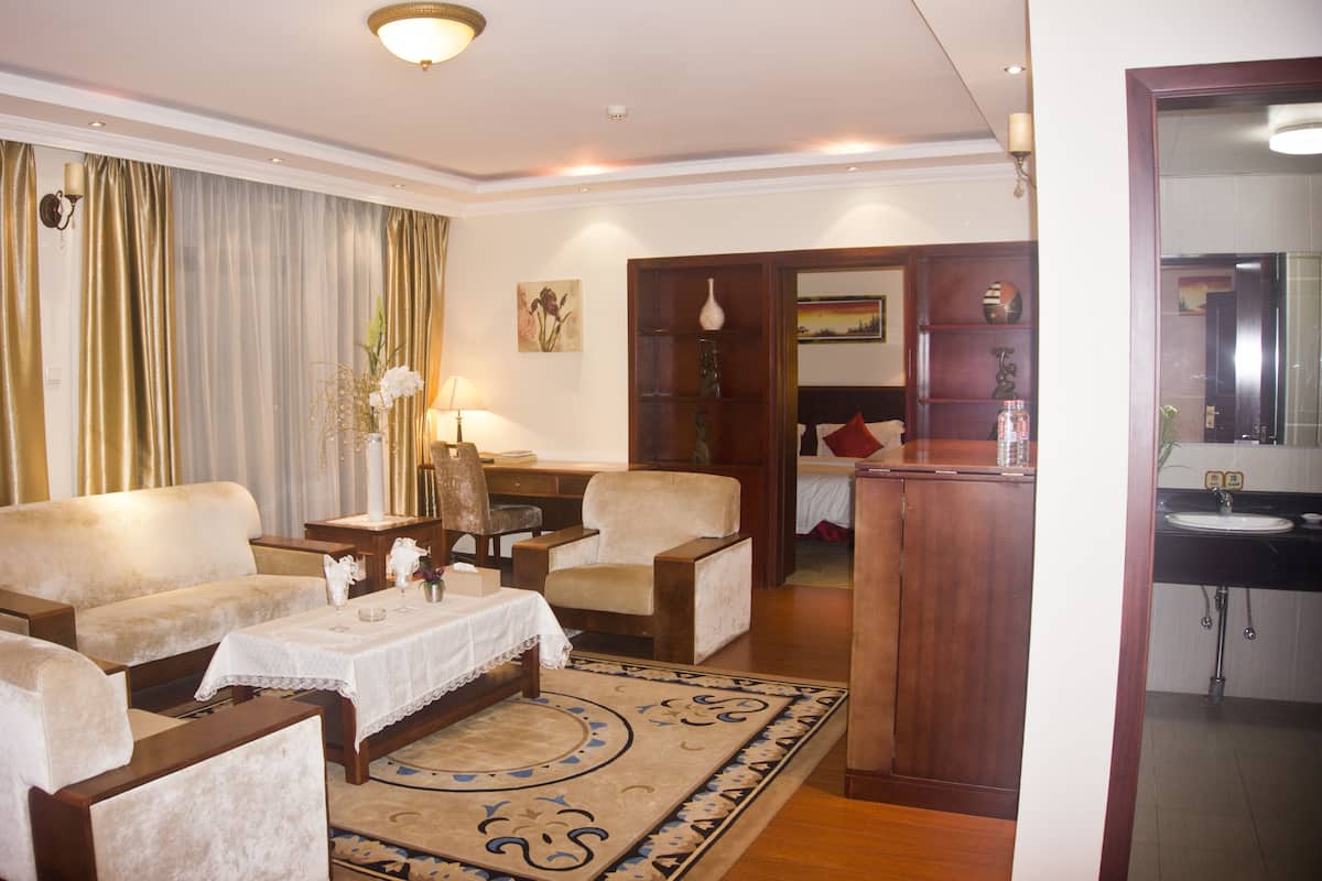A hotel room with a living area featuring a sofa, armchairs, a coffee table, and a dining table. There is a bathroom visible through an open door.