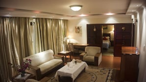 A hotel room with a sofa, armchair, small table, and a desk with a chair. There is a bed in the background and a wardrobe. - GS Plaza Hotel (Accra)