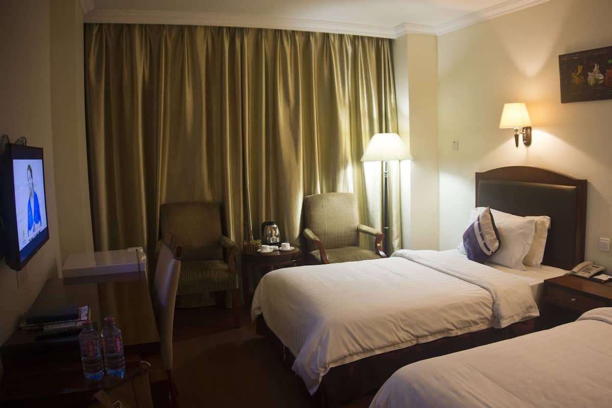 A hotel room with two beds, a chair, a desk with a computer, a lamp, and a painting on the wall.