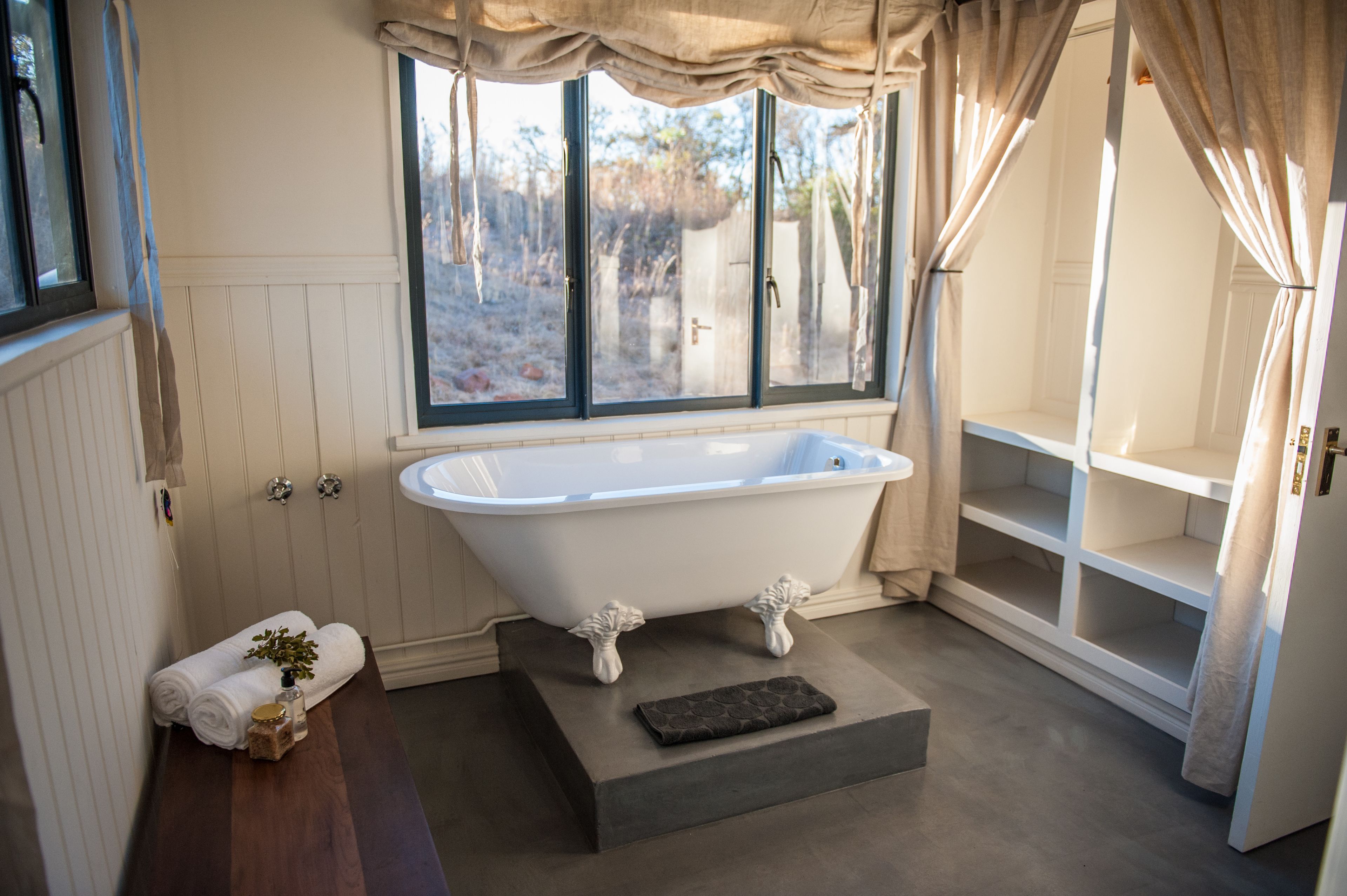 Un baño con una bañera independiente, una ventana con cortinas, un banco de madera y un perchero.