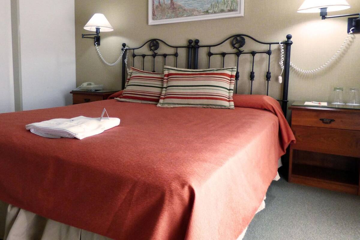 A neatly made bed with a red bedspread and striped pillows, a bedside table with a lamp, and a telephone.