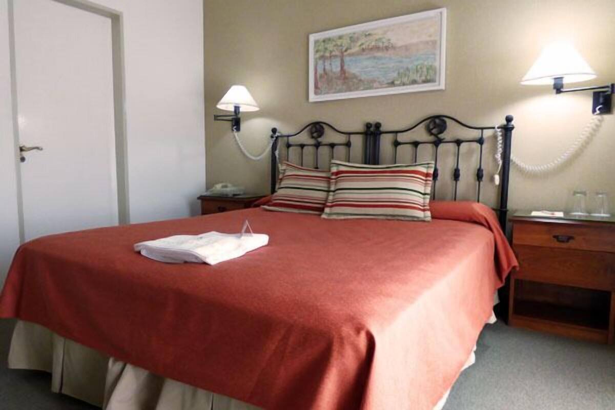 A neatly made bed with a red bedspread, two pillows, a white towel, and a bedside table with a lamp and a phone.