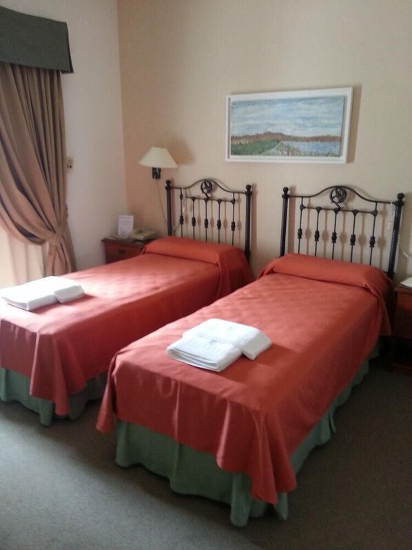 A hotel room with two single beds, each with a red bedspread and green skirted footboard. There is a painting on the wall above the beds, a lamp on the nightstand, and a window with curtains.