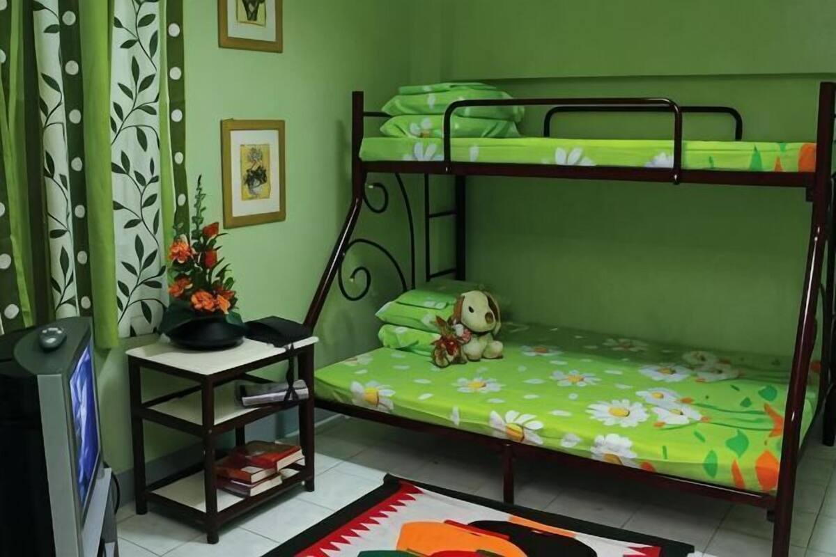 Una cama litera con una colcha estampada de flores verdes, una mesita con una planta y un cuadro en la pared.
