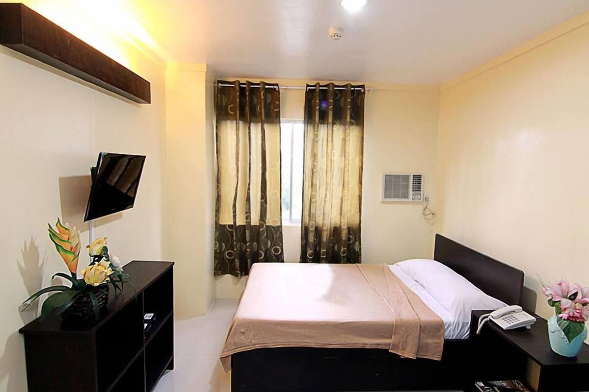 Habitación de hotel con una cama, un televisor sobre un mueble, un teléfono y una pequeña mesa con un florero.