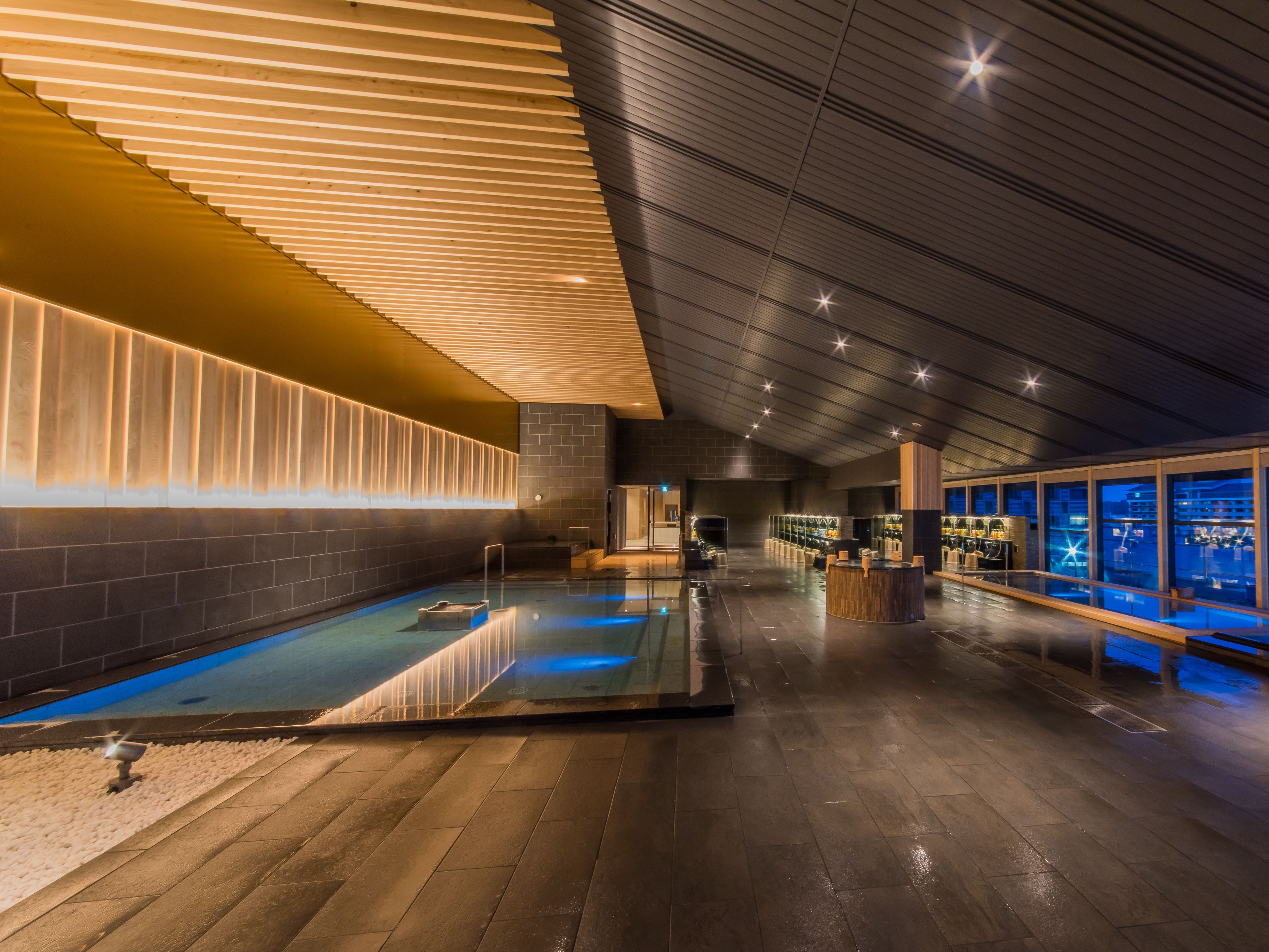 A modern indoor pool area with a wooden floor, a pool with a blue tint, and a bar area with a view of the city.